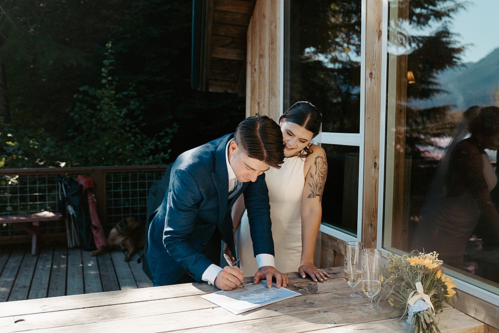 Groom signing a marriage license on a picnic table with bride next to him.