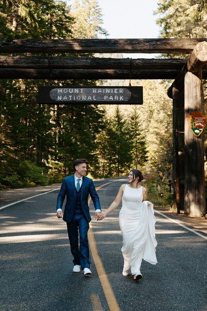 Bride and groom holding hands while walking in the middle of the road with the Mount Rainier National Park sign behind them.