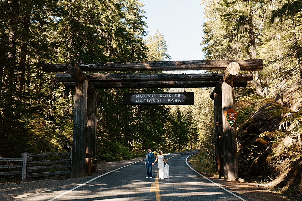 Bride and groom holding hands and looking in opposite directions while standing in the road under the Mount Rainier National Park sign.