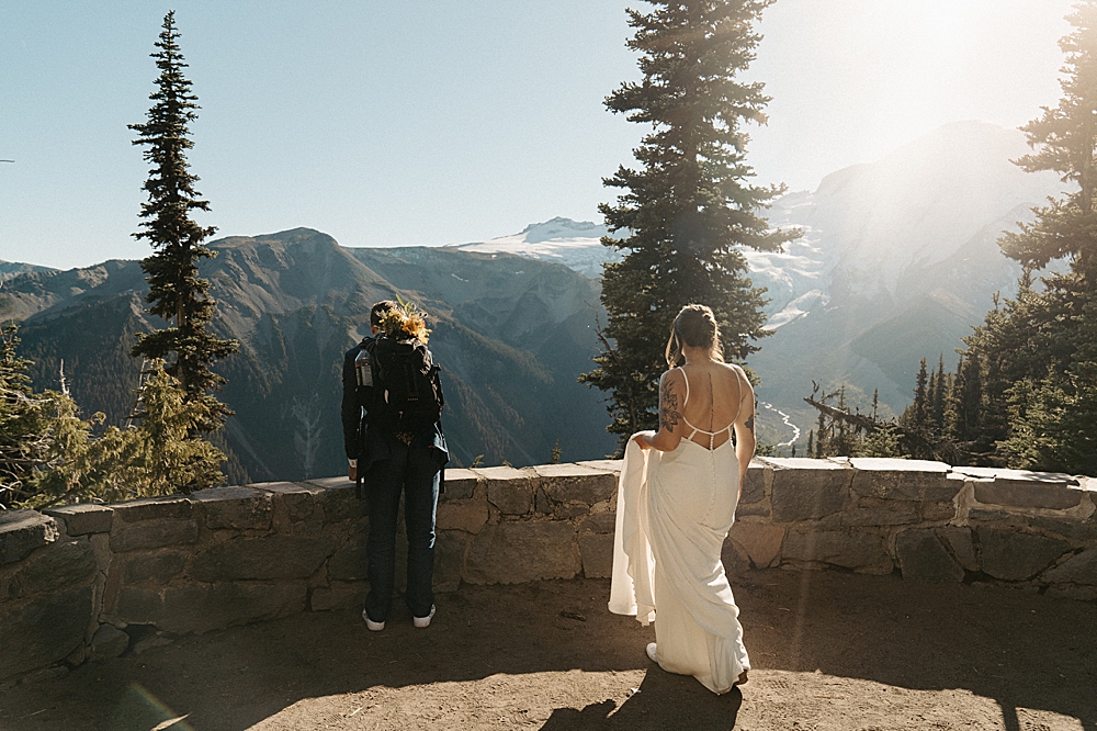 Groom standing and looking over a short rock wall with the bride holding her train and walking towards him at an overlook in Mount Rainier National Park.