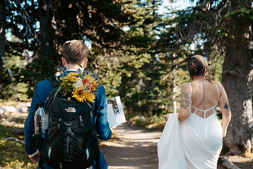 Groom in a navy blue suit walking on a trail with a floral bouquet in his hiking pack and a cake box in his hands with the bride holding her train and walking next to him.