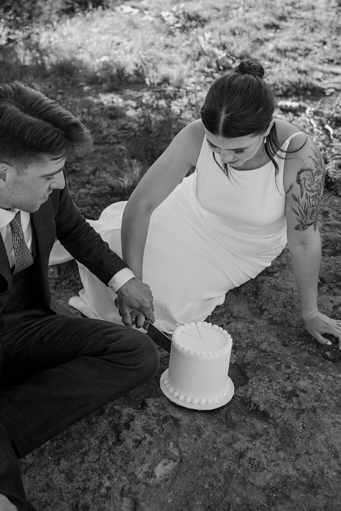 Black and white of bride and groom sitting on a rock while cutting a small, white wedding cake.