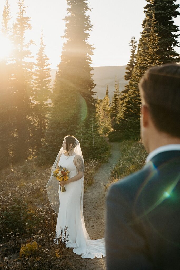 Groom blurry in the foreground with his attention on the bride holding a floral bouquet and standing on a trail in Mount Rainier at sunset.