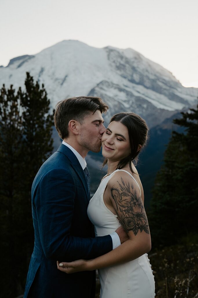 Groom kissing his bride's cheek with Mount Rainier behind them.
