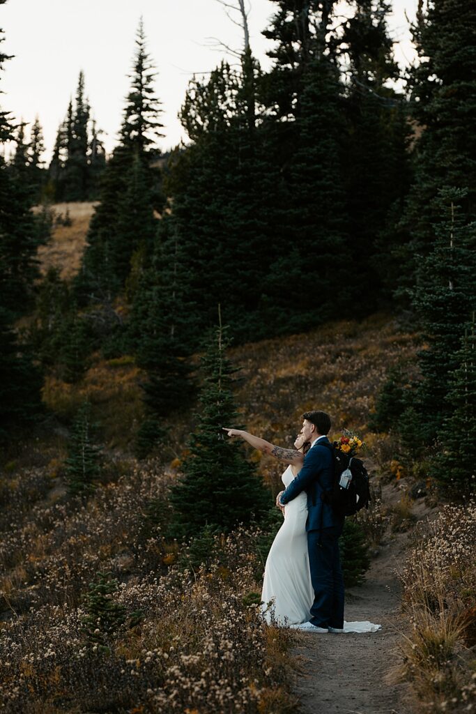 Groom with a hiking pack and floral bouquet holding onto his bride while she points at the mountains.