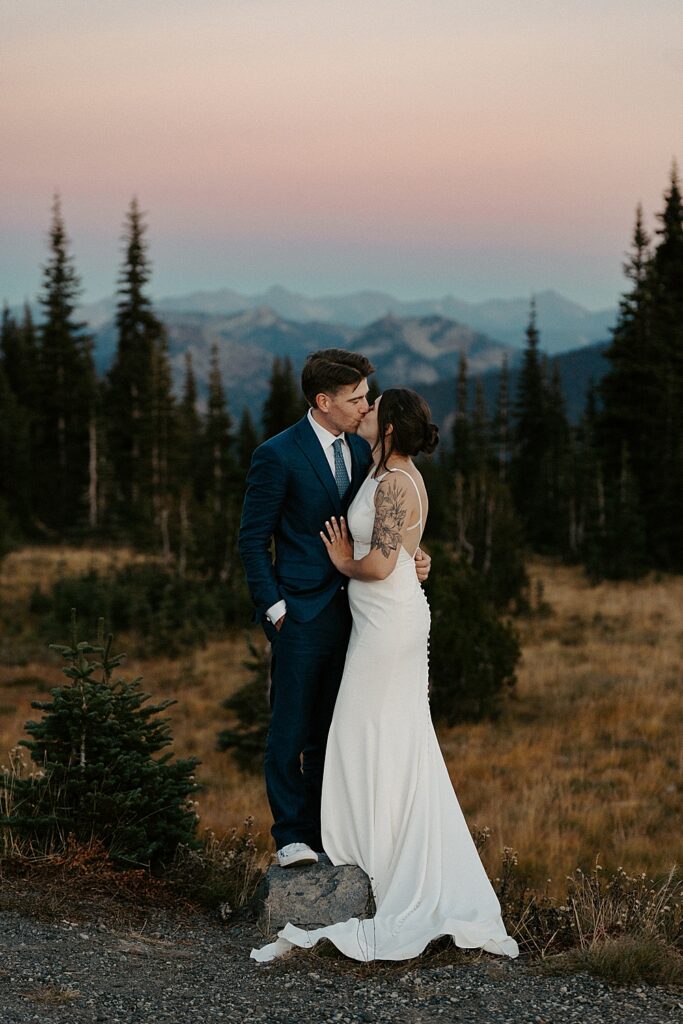 Bride and groom kissing while standing on a rock after the sun has set and the sky is colorful.