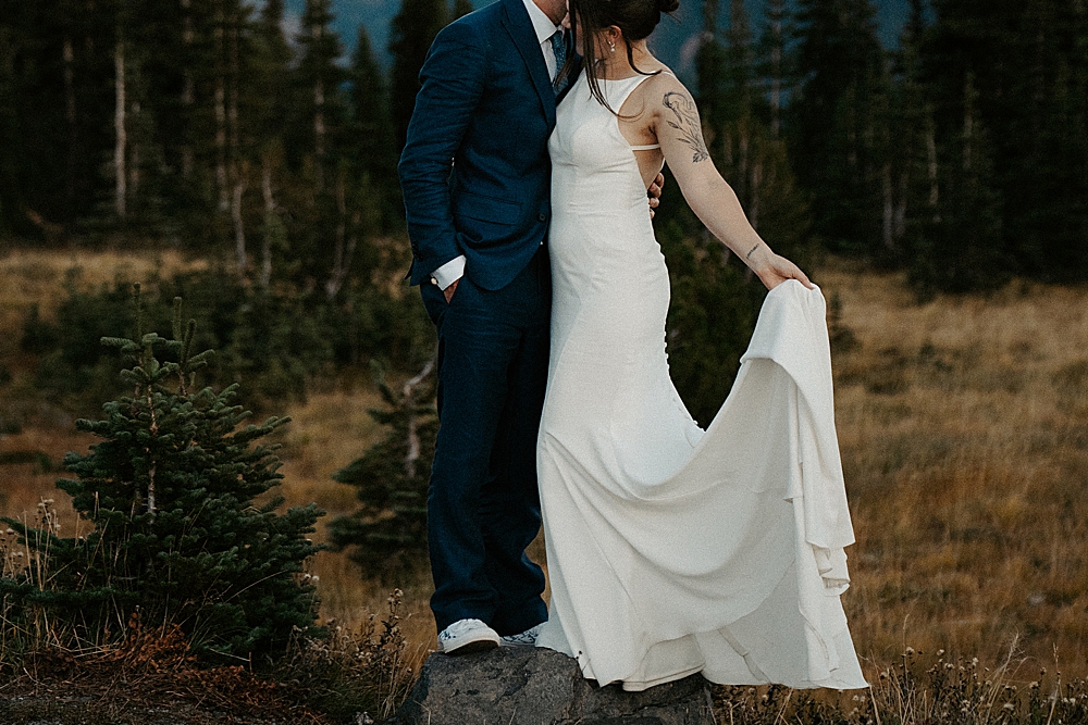 Up close of bride holding her train while leaning into the groom and standing on a rock. 