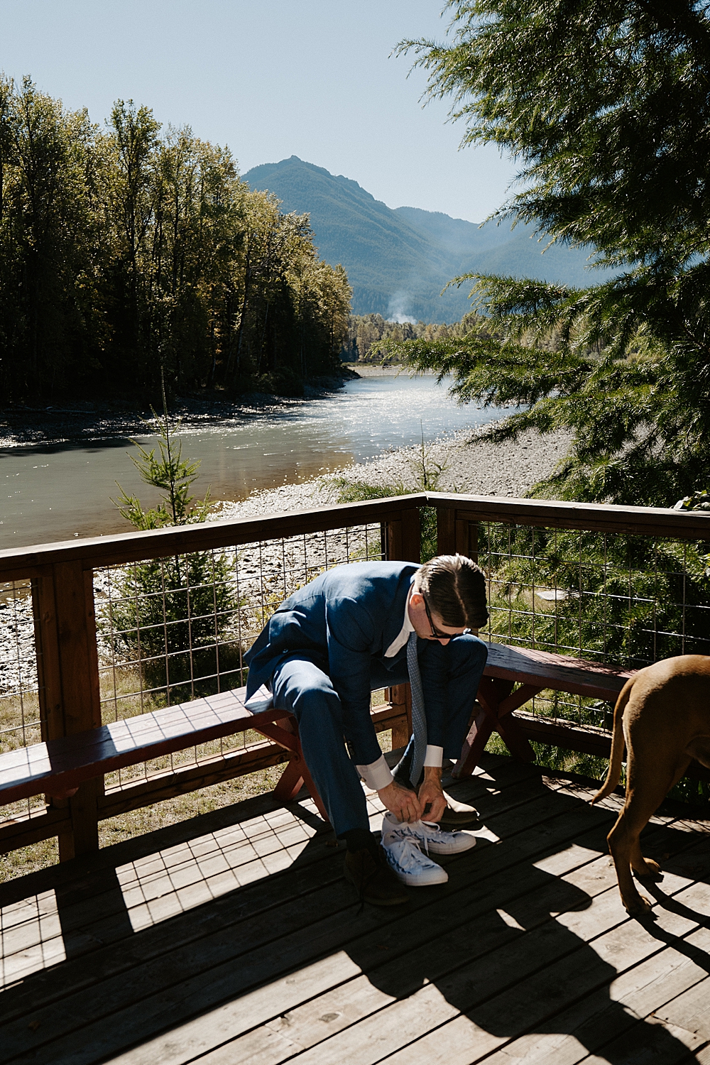 Groom putting shoes on while sitting at a bench on a cabin's porch with a river and mountains in the background.