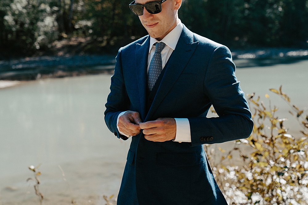 Groom buttoning his navy blue suit jacket with sunglasses on.