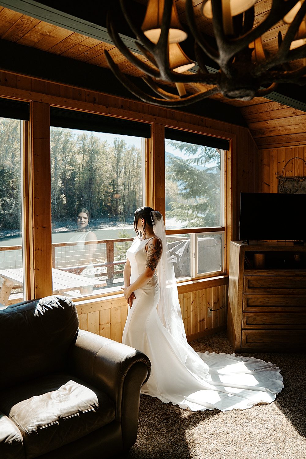 Bride in a satin wedding dress with a long veil standing in front of large windows in a cozy cabin.