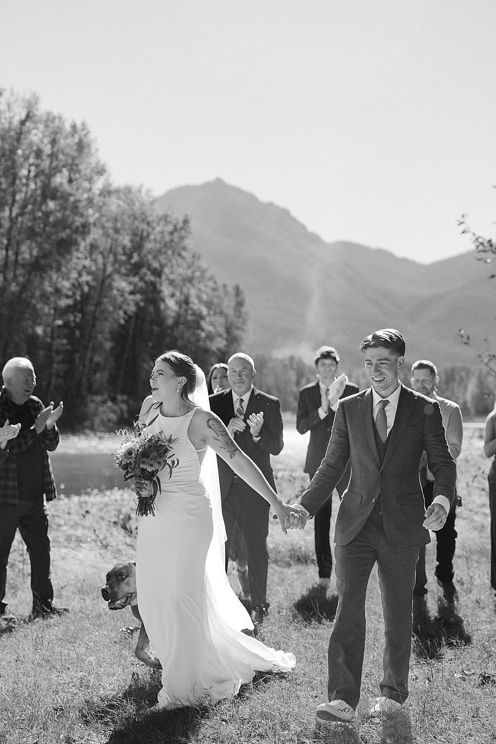 Black & white of bride and groom holding hands and walking on a lawn after their ceremony with family walking behind them.