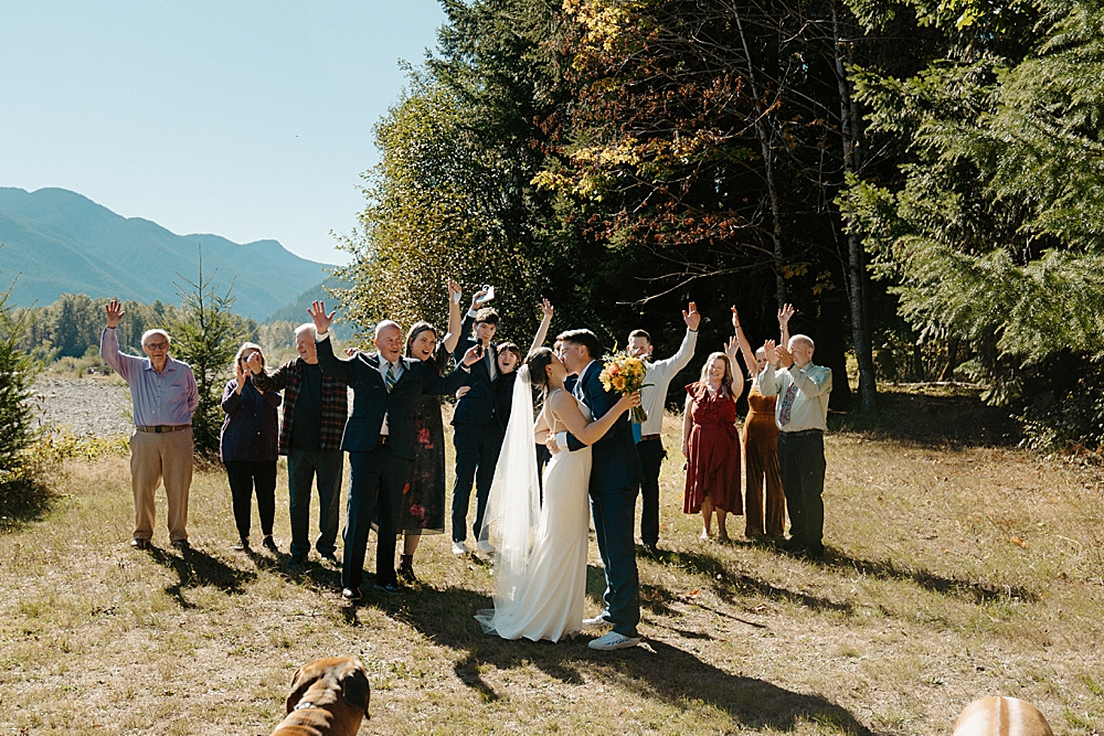 Bride and groom kissing with all of their family cheering behind them.