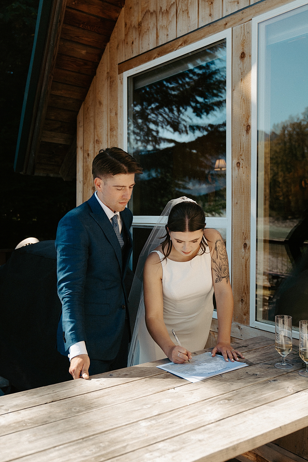 Bride signing marriage license on a picnic table with her groom standing next to her.
