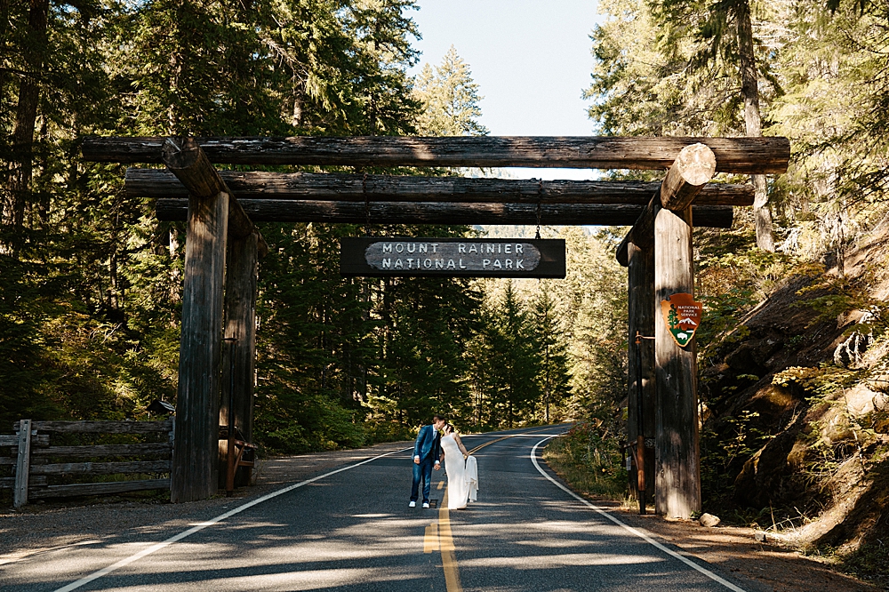 Bride and groom standing under the Mount Rainier National Park sign in the road and kissing.