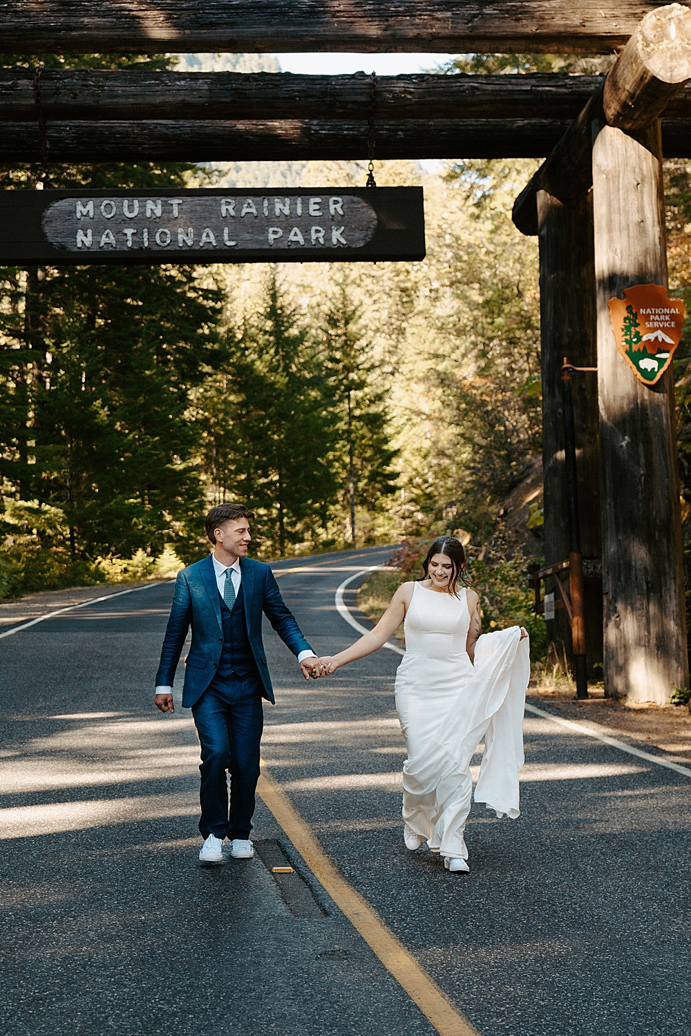Bride in a satin dress and groom in a navy suit holding hands and walking in the road under the Mount Rainier National Park sign.