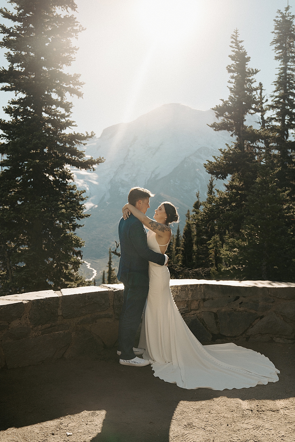 Bride and groom standing at an overlook in Mount Rainier with the mountain and sunshine behind them.