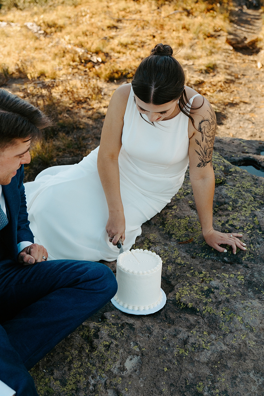Bride and groom sitting on a rock and cutting into a white wedding cake with "just married" written on the top.