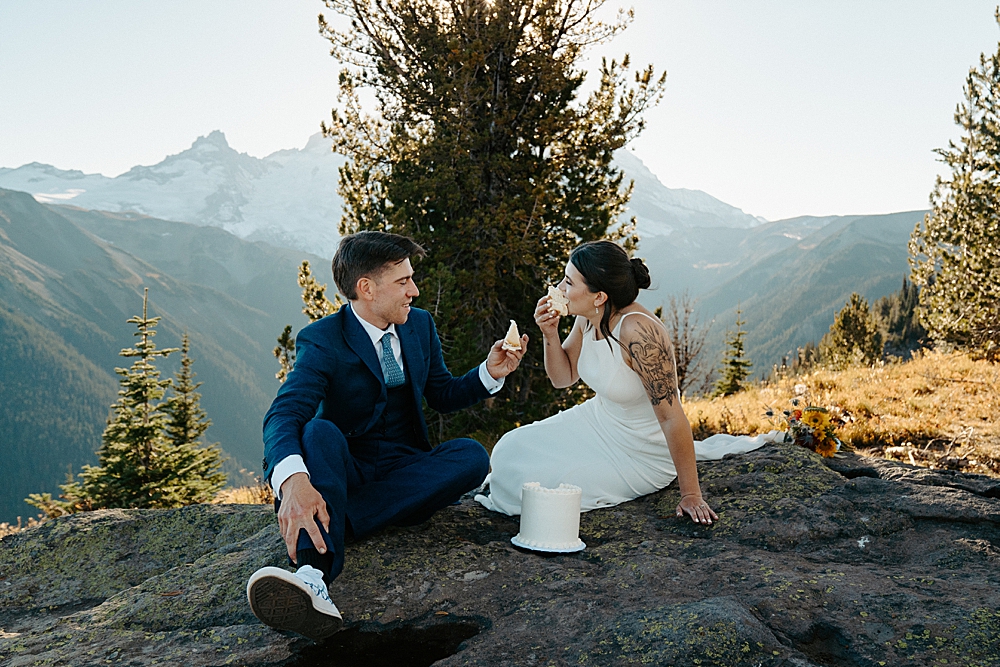 Bride and groom sitting on a rock in Mount Rainier National Park with slices of wedding cake in their hands.