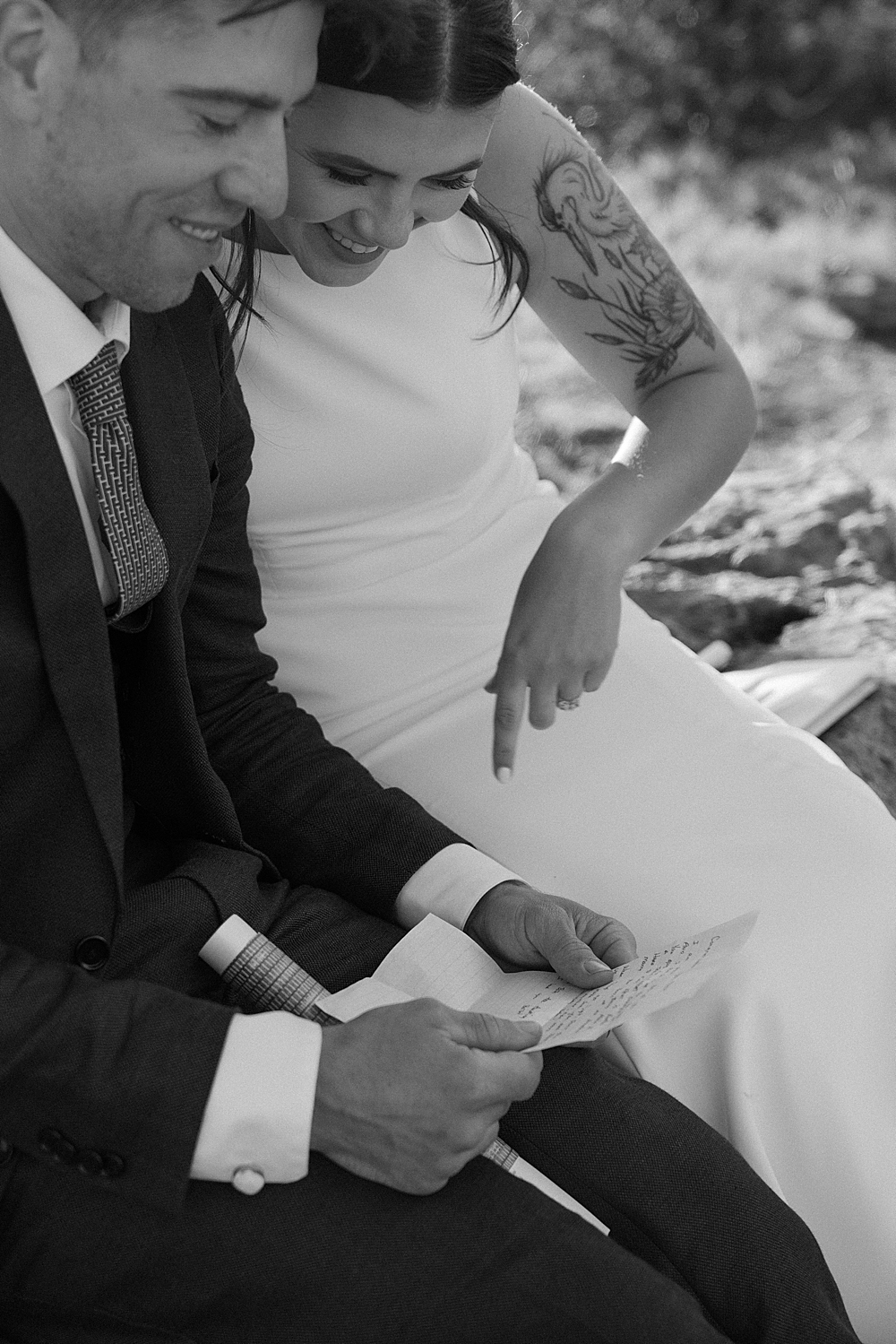 Black & white of a bride and groom sitting on a rock while reading a letter from a loved one.