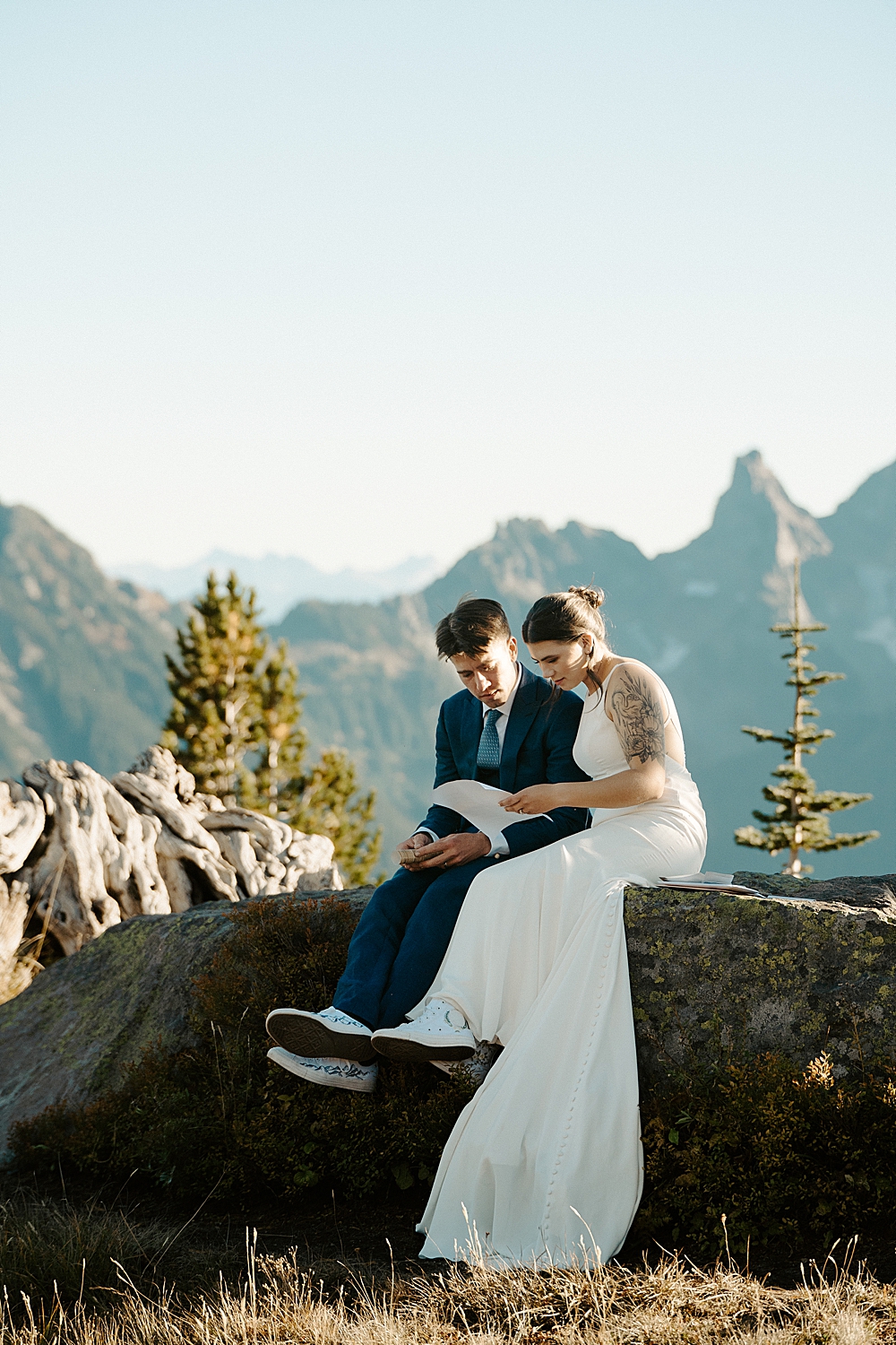 Bride in a satin dress sitting on a rock next to her groom in a navy blue suit while reading a letter.