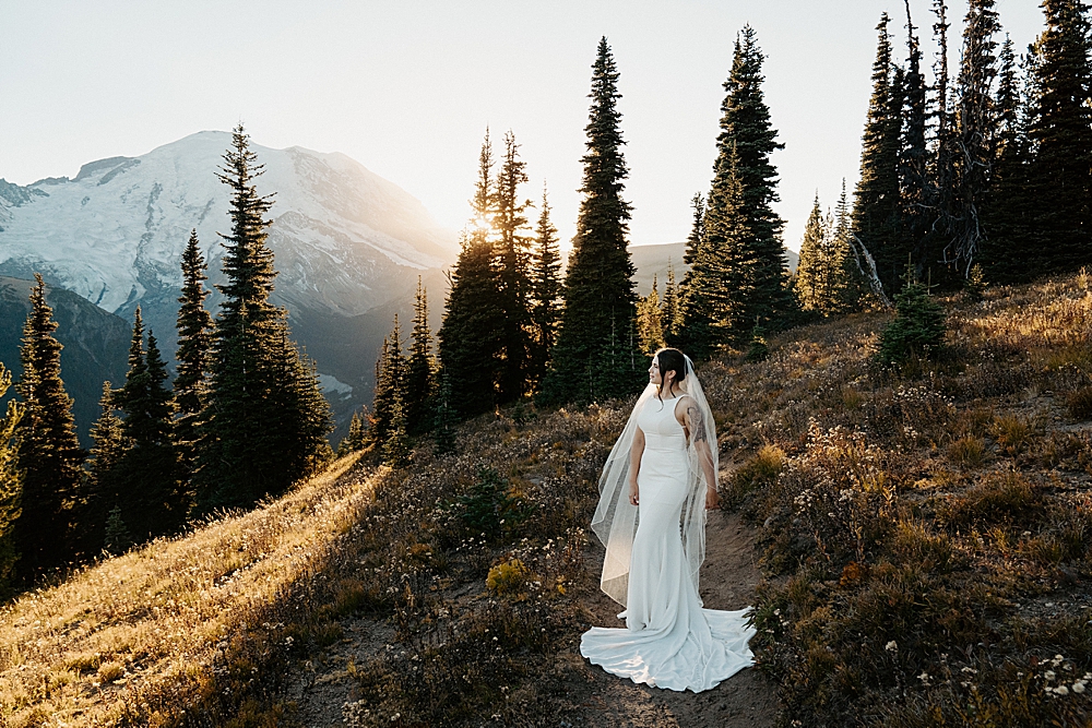 Bridal portrait of a woman in a satin dress and a long veil standing on a trail in Mount Rainier National Park at sunset.