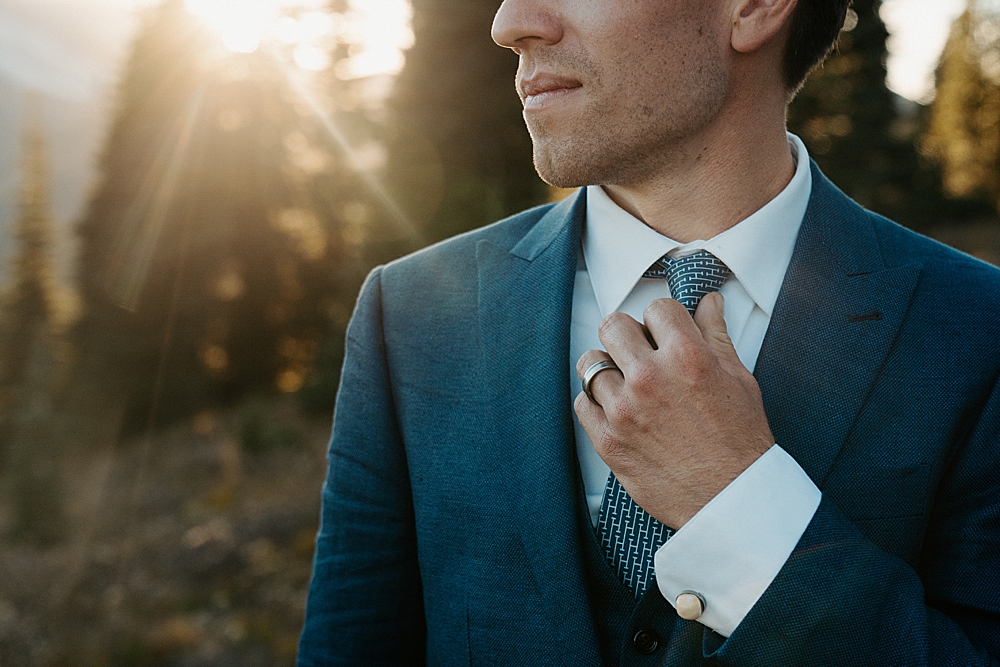 Up close of groom in a navy blue suit adjusting his blue and white patterned tie at sunset.