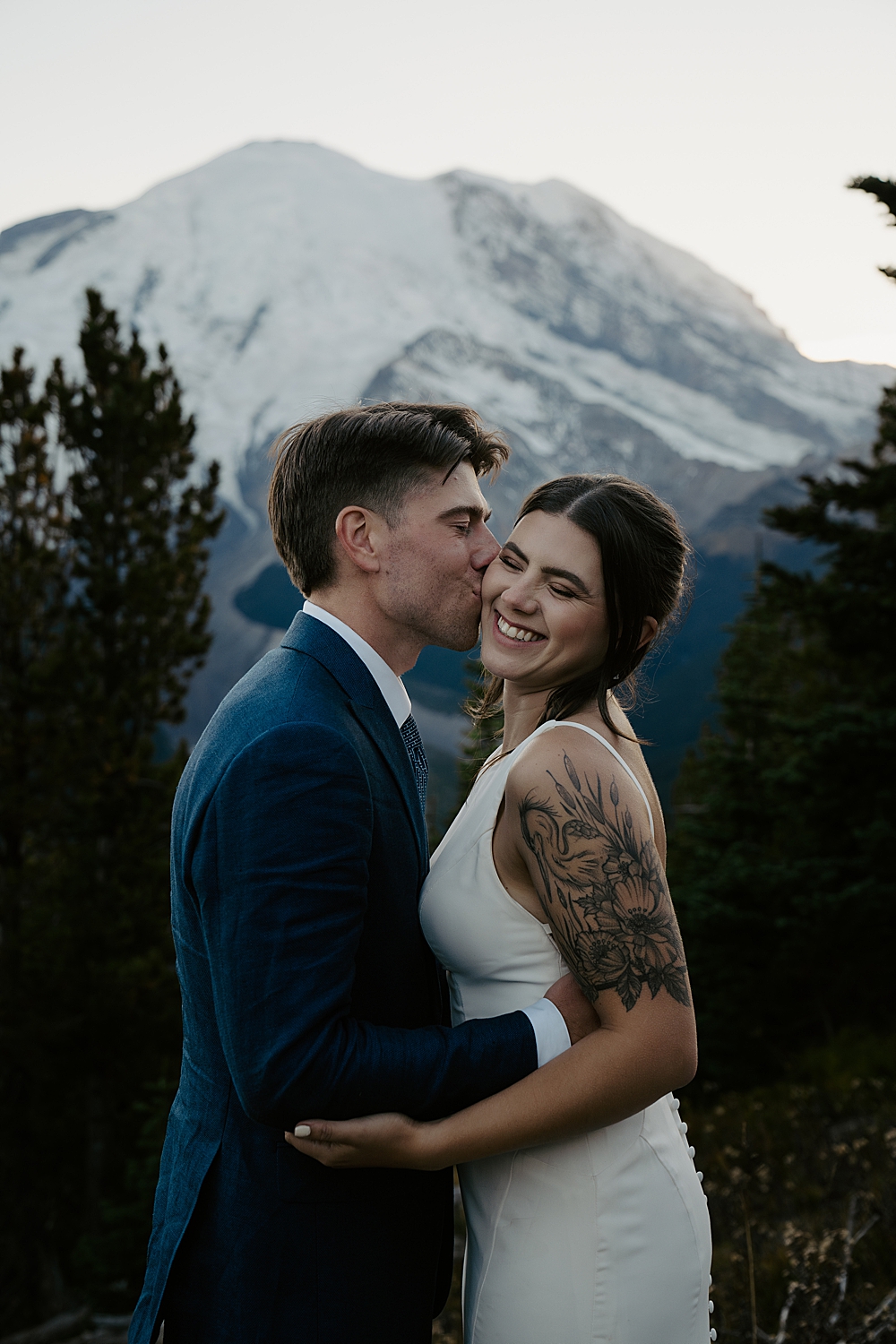 Groom in a navy suit kissing his bride on the cheek while she's smiling with Mount Rainier in the background after the sun has set.