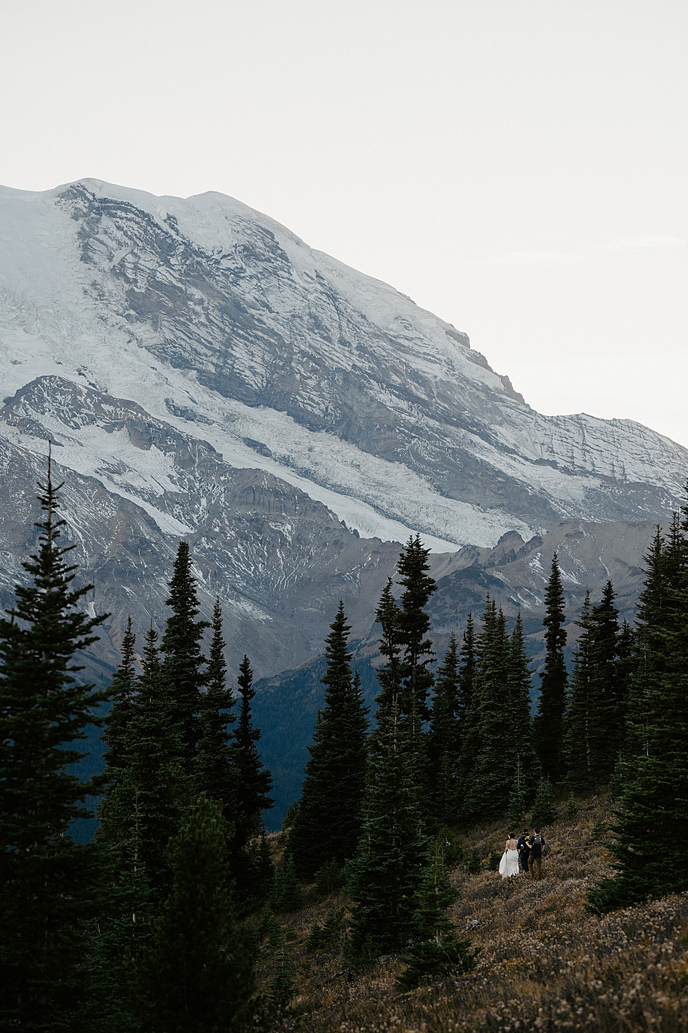 far away photo of bride, groom, and videographer from Venture to Elope hiking on a trail with Mount Rainier as the focus.