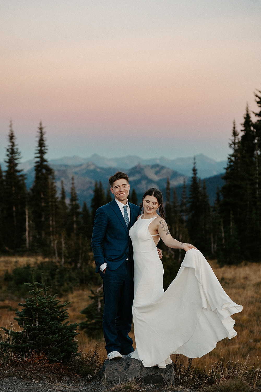 Bride holding her wedding dress train and standing next to her groom in a navy blue suit with a colorful sky after sunset behind them.