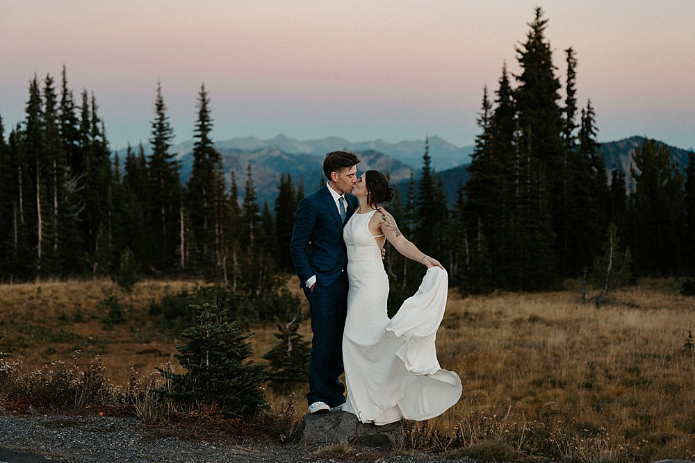 Bride and groom kissing while standing on a rock after sunset with a colorful sky in the background.