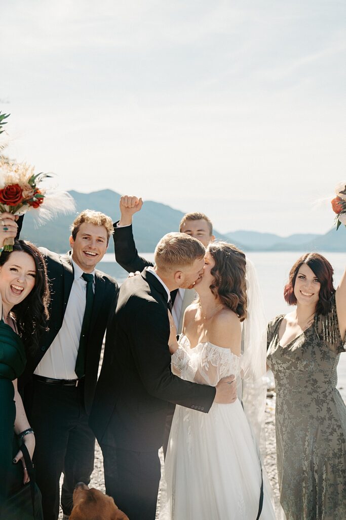 Bride and groom kissing at Lake McDonald surrounded by groomsmen and bridesmaids cheering 