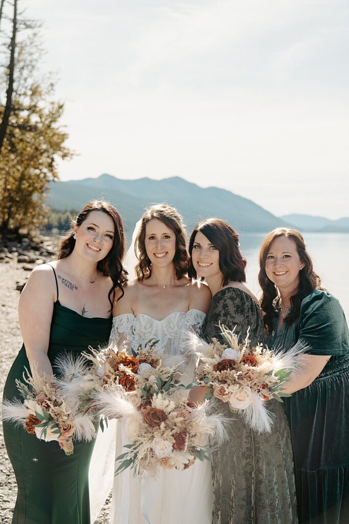 Bride with a dried fall bouquet and three bridesmaids in forest green dresses smiling along Lake McDonald Beach.