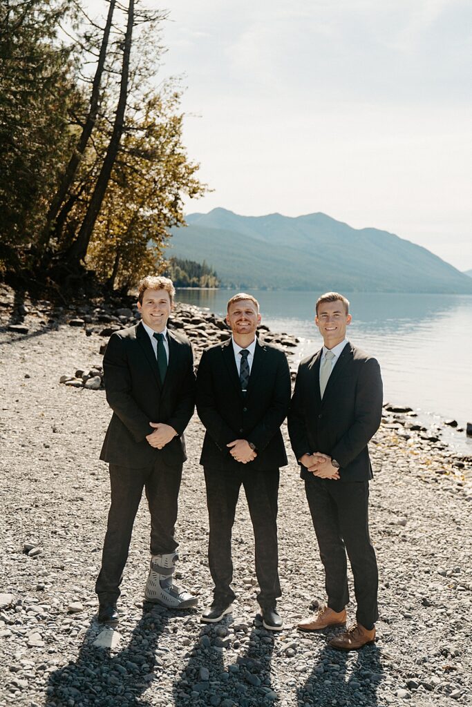 Groom in a black wedding suit with two groomsmen smiling along the shore of Lake McDonald in Glacier