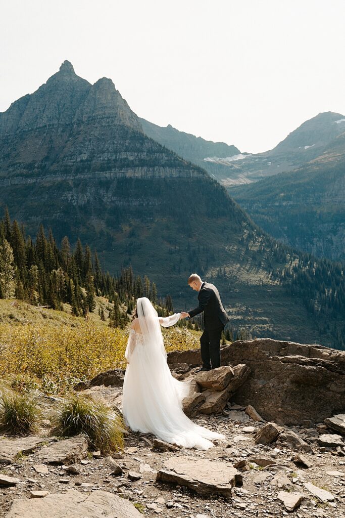 Groom in a black suit helping bride up a rock at Big Bend in Glacier Park