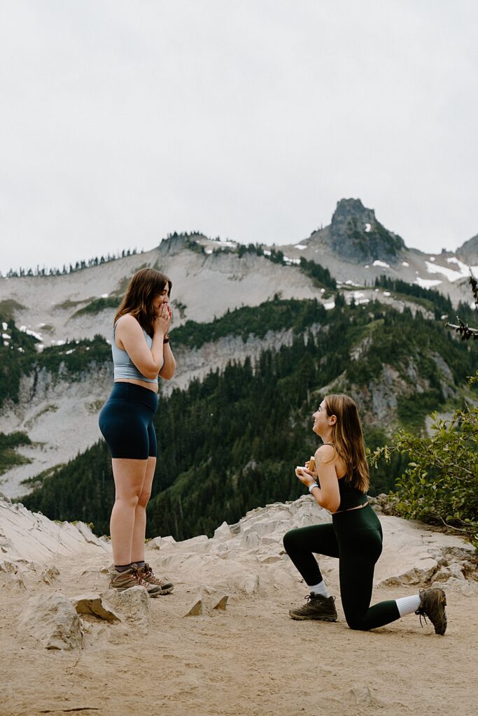 One girl in hiking attire proposing to her girlfriend on a hiking trail at Mount Rainier.