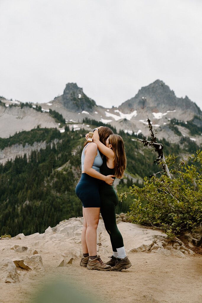 Two girls in hiking attire hugging and kissing after their surprise proposal in Mount Rainier National Park.