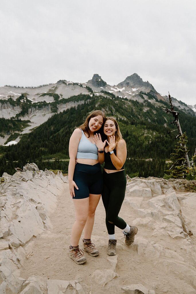 Two girls in hiking attire smiling and showing their rings after proposing to each other in Mount Rainier National Park.