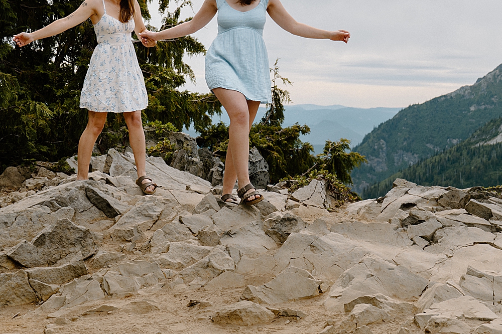 Two girls holding hands in dresses and sandals walking on rocks after their Mount Rainier proposal.
