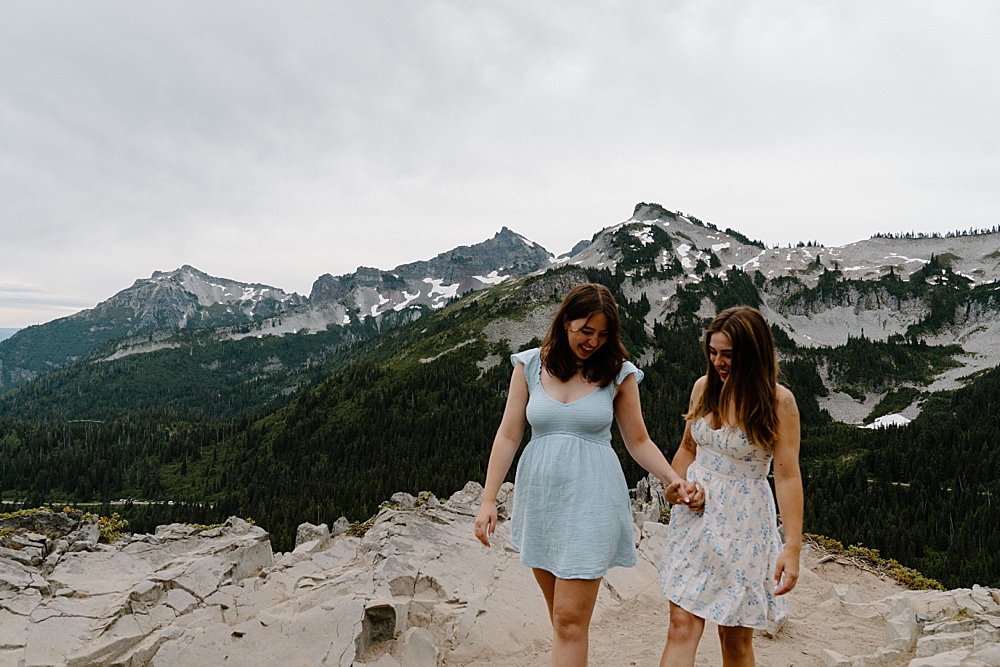 Two girls in dresses holding hands and walking together after proposing to each other with views of the Tatoosh Mountains Range in Washington.
