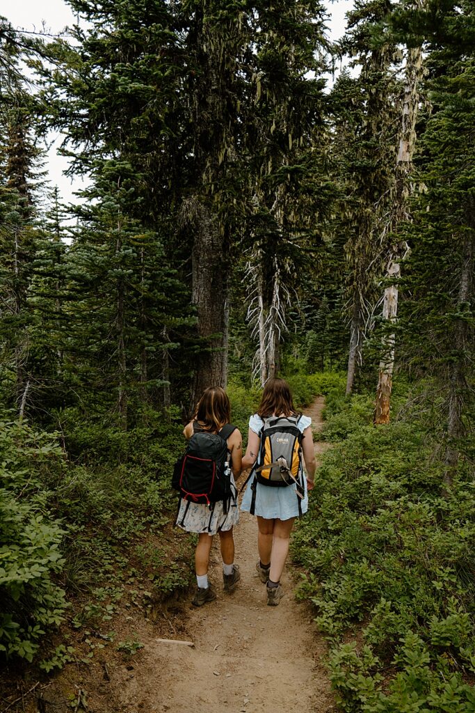 Two girls in dresses, hiking boots, and backpacks holding hands and walking on a Mount Rainier trail.