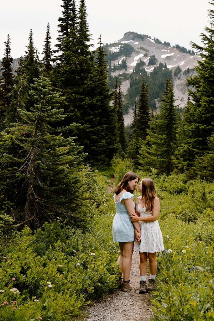 Two girls in dresses and hiking boots holding each other on a trail at Mount Rainier after their double surprise proposal during wildflower season.