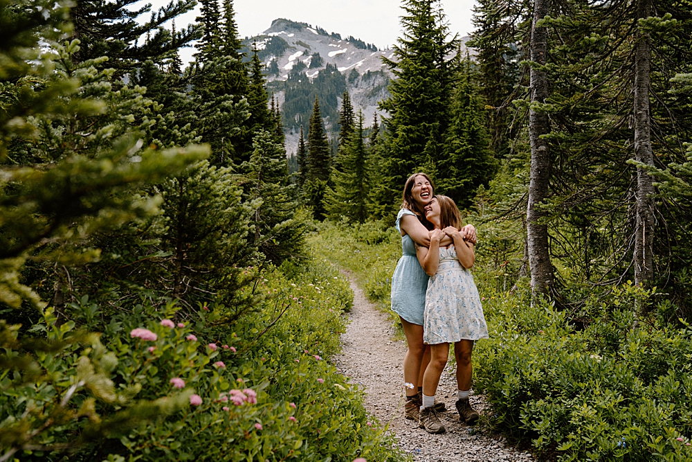 Two girls in dresses and hiking boots hugging and laughing on a trail at Mount Rainier after their double surprise proposal during wildflower season.