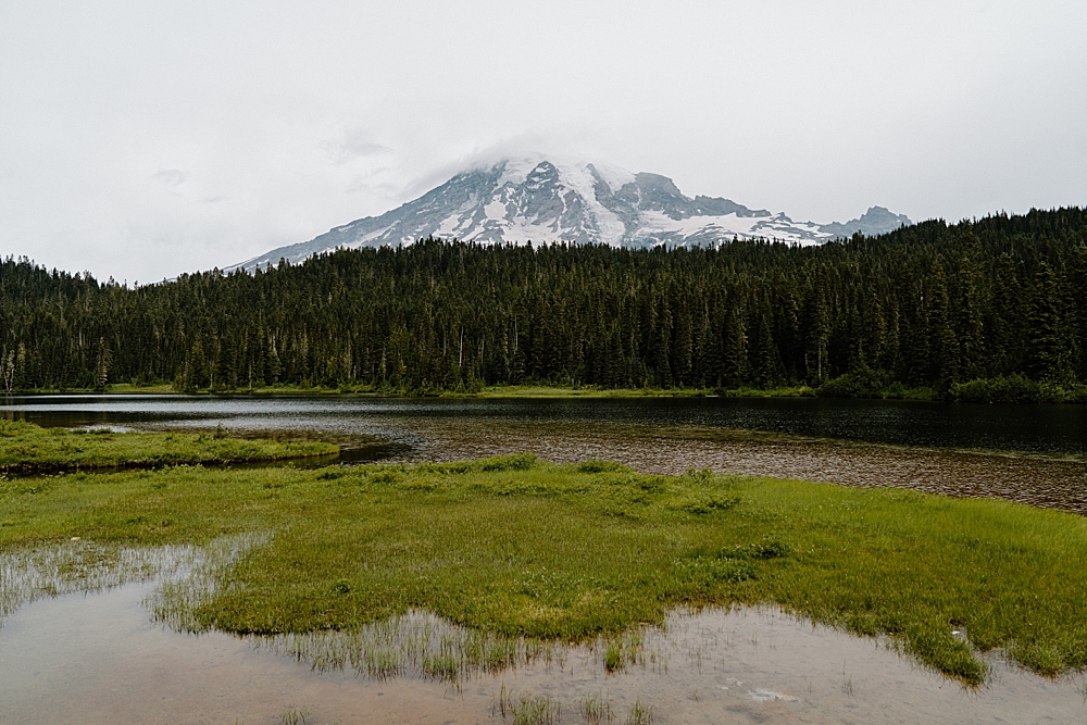 Clouds slightly covering the top of Mount Rainier with trees and Reflection Lake in the foreground.