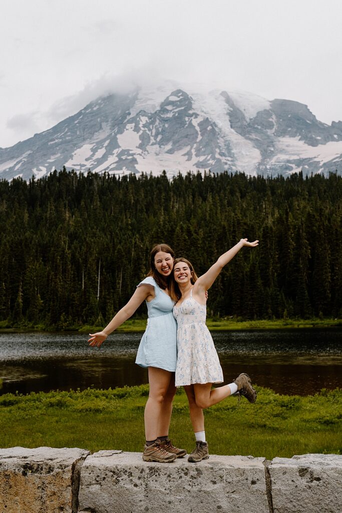 Two girls in hiking boots and dresses standing on a rock wall cheering after their proposal with Mount Rainier in the background.