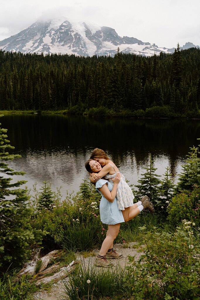 One girl in a short, blue dress and hiking boots holding and lifting her fiancée who is also in hiking boot and a dress with Mount Rainier in the background.