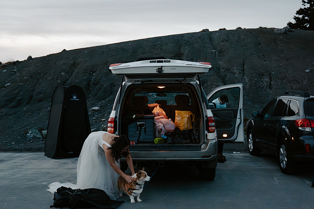 Bride in a wedding dress and corgi standing behind an SUV before sunrise getting ready for her elopement. 