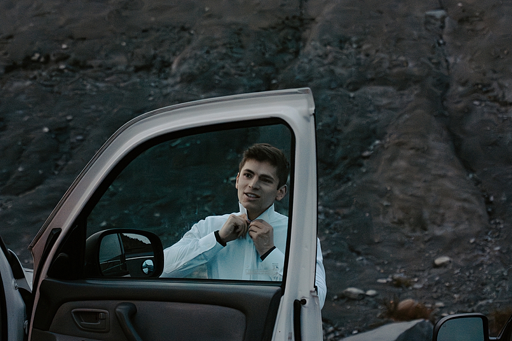 Groom buttoning his dress shirt using the reflection of a car window before his sunrise elopement.