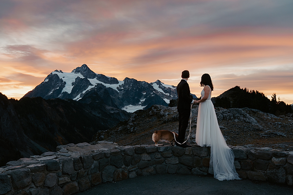 Bride, groom, and corgi standing on a low rock wall facing sunrise and Mount Shuksan while holding hands.
