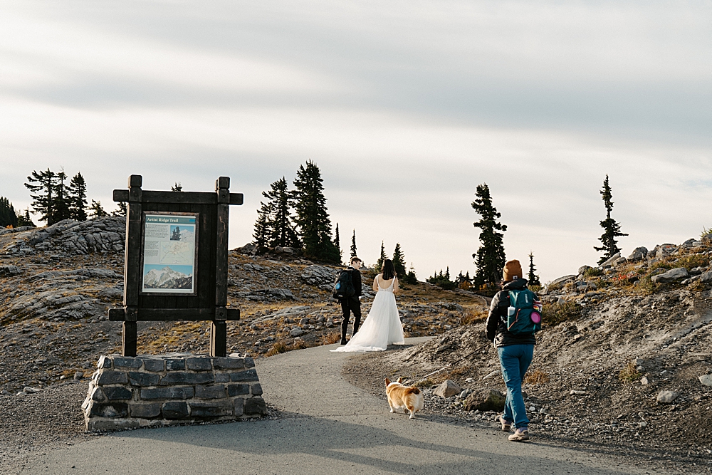Bride and groom walking along the Artist Ridge Trail with dog-handler and corgi walking behind them.