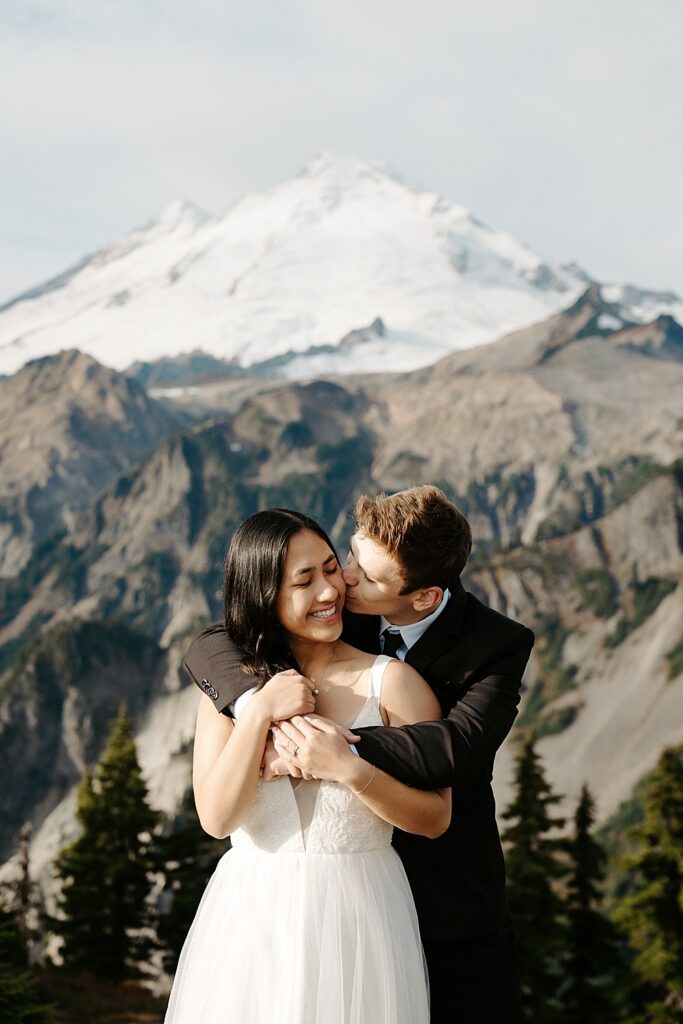 Groom kissing bride's cheek with his arms wrapped around her and Mount Baker in the background. 